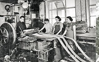 Historisches Foto: Mitarbeitende arbeiten gemeinsam an einer Maschine zur Verarbeitung von Vorhangschienen.