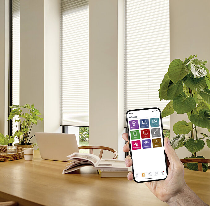 Hand mit Smartphone, das die PowerView® App zeigt, im Wohnraum mit MHZ Wabenplissees an Fenstern.