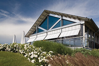 Gebäude mit grosser Glasfassade, ausgestattet mit weiss-grau gestreiften ZIP-Senkrechtmarkisen, davor bepflanzte Grünfläche.