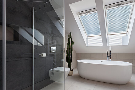 Badezimmer mit freistehender Wanne und Jalousien im Dachfenster. Lamellen verlaufen parallel zur Fensterfläche und filtern das Licht.