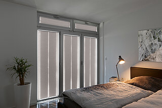 Lichtundurchlässige graue Plissees an bodentiefen Fenstern in einem modernen Schlafzimmer.