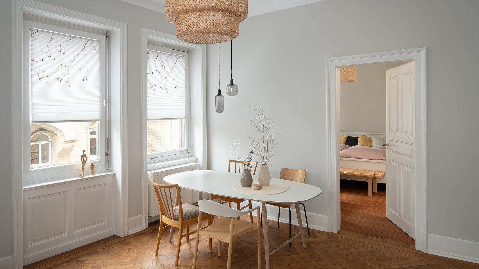 Zwei weisse Fenster mit floralen MHZ-Wabenplissees im hellen Esszimmer mit hölzernem Fischgrätboden.