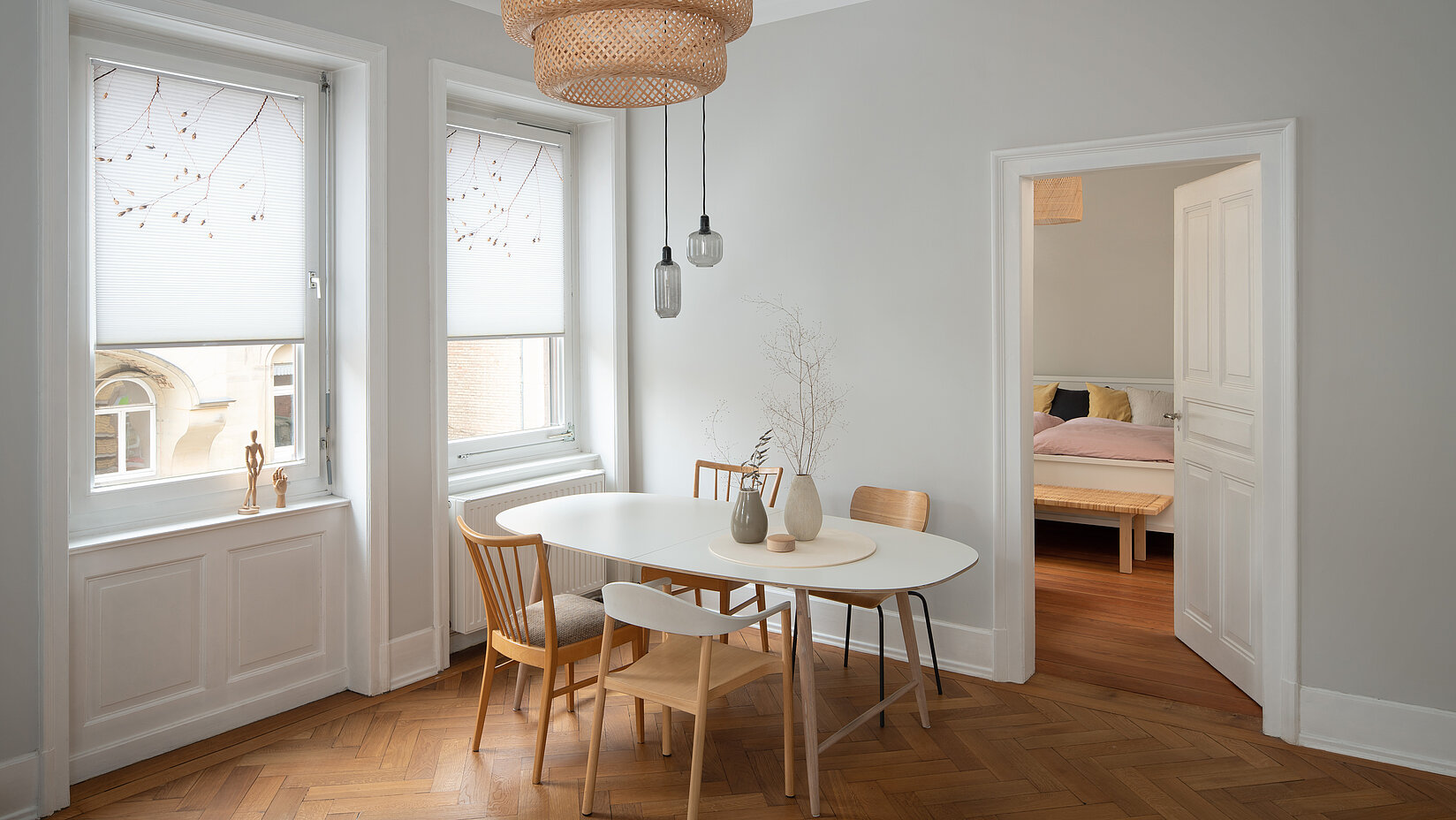 Zwei weisse Fenster mit floralen MHZ-Wabenplissees im hellen Esszimmer mit hölzernem Fischgrätboden.