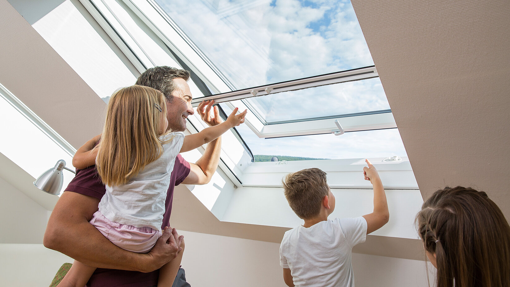 Familie bedient Insektenschutz-Rollo an einem geöffneten Dachfenster im Dachgeschoss.