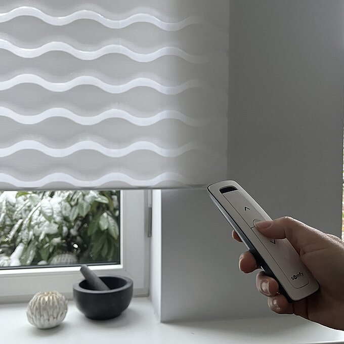 Hand mit Somfy-Fernbedienung steuert das MHZ Doppelrollo „Loki“ mit weissem Wellenmuster an einem Fenster.