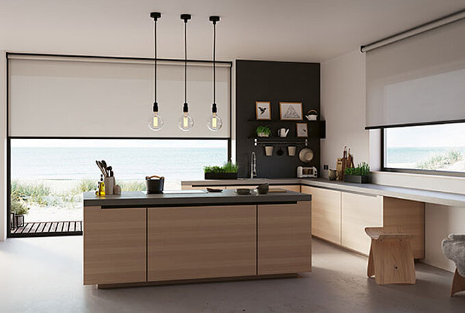 Moderne Küche mit grosser Kochinsel und zwei Fenstern mit hellen Rollos, Blick auf Dünenlandschaft am Meer.