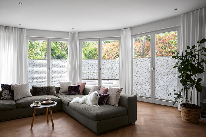 Wohnzimmer mit weissen floralen Plissees als halbtransparenter Sichtschutz an bodentiefen Fenstern.
