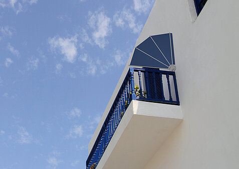 Balkon mit blauer Metallbrüstung und seitlich montierter fächerförmiger Markise an weisser Fassade