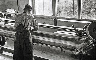 Ein Mann in Schürze arbeitet an einer Drehmaschine zur Bearbeitung eines langen Werkstücks.