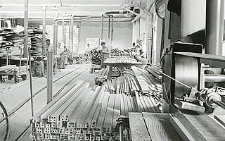 Historisches Schwarz-Weiss-Foto einer Werkshalle mit Mitarbeitenden bei der Fertigung von Vorhangschienen.