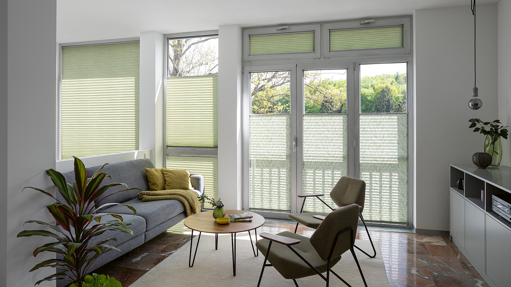 Grüne Plissees mit einfarbigen und floralen Bereichen in hellem Wohnzimmer mit bodentiefen Fenstern.