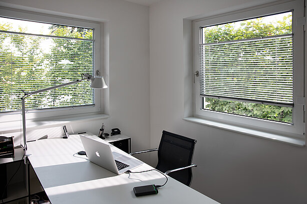 Homeoffice mit zwei Fenstern und silberfarbenen Jalousien. Lamellen regulieren das Licht für blendfreies Arbeiten.