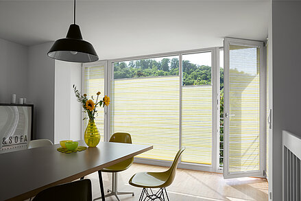 Plissee „Manolya“ mit gelb-beigem Streifenmuster in hellem Essbereich mit gelben Stühlen.