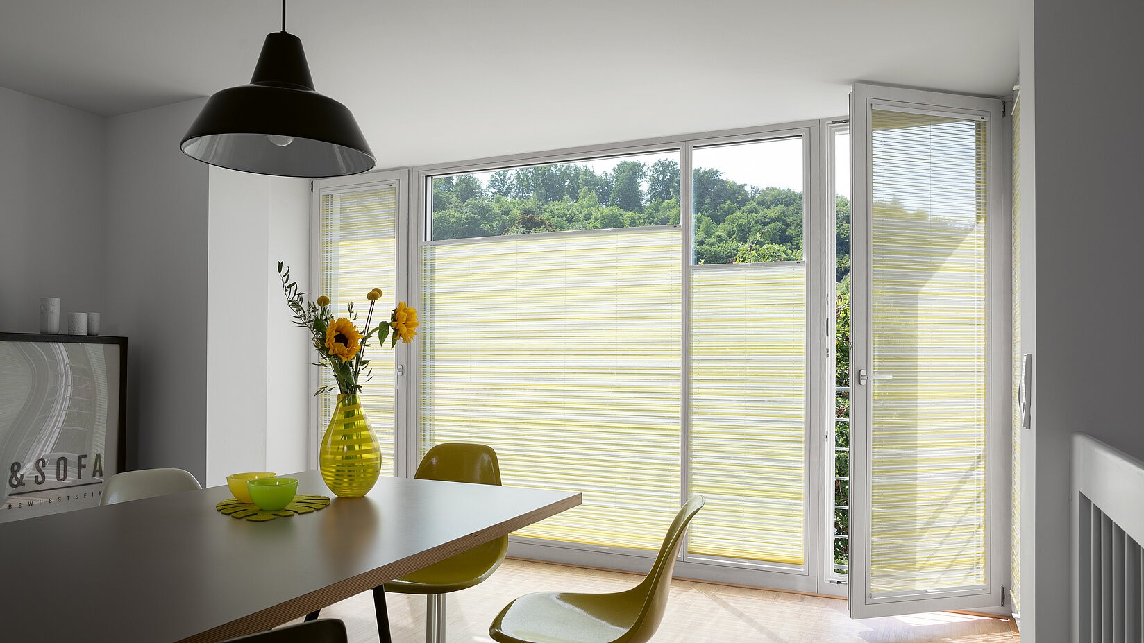 Plissee „Manolya“ mit gelb-beigem Streifenmuster in hellem Essbereich mit gelben Stühlen.