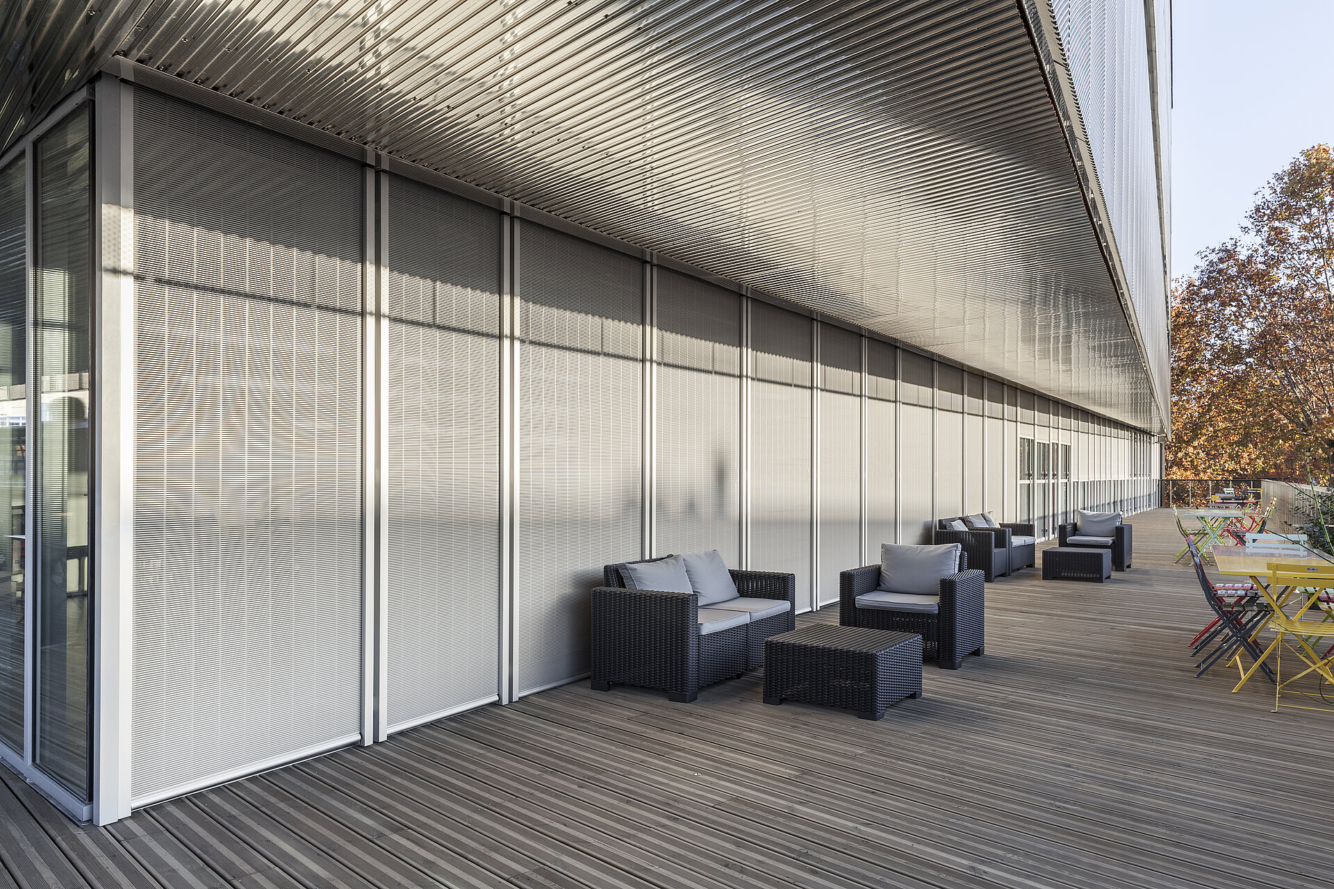 Terrasse mit Sitzmöbeln vor einer Glasfassade mit aussenliegenden vertikalen Senro Metallbehängen als Sonnenschutz.