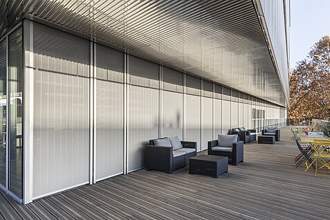 Terrasse mit Sitzmöbeln vor einer Glasfassade mit aussenliegenden vertikalen Senro Metallbehängen als Sonnenschutz.