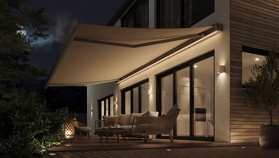 Beige Gelenkarmmarkise mit integrierter Beleuchtung über Terrasse eines Hauses bei Abenddämmerung.