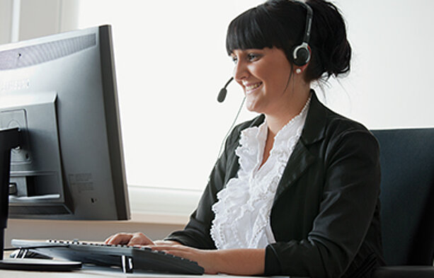 Frau mit Headset sitzt vor einem Monitor und spricht freundlich mit einem Kunden.