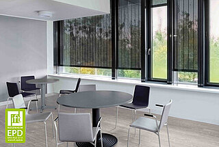 Ein heller Aufenthaltsraum mit mehreren Stühlen und runden Tischen. An den grossen Fenstern sind schwarze vertikale Metallbehänge montiert, die als Sicht- und Sonnenschutz dienen.