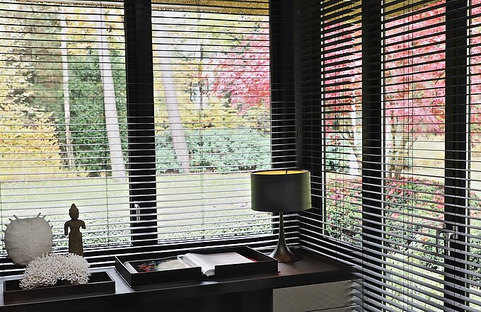 Schwarze Innenjalousien vor Fenstern mit Blick auf bunten Garten, Sideboard mit Deko im Vordergrund.