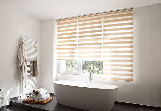 Modernes Badezimmer mit freistehender Badewanne und MHZ Doppelrollo „Alva“ in Beige, das Licht filtert und Sichtschutz bietet.