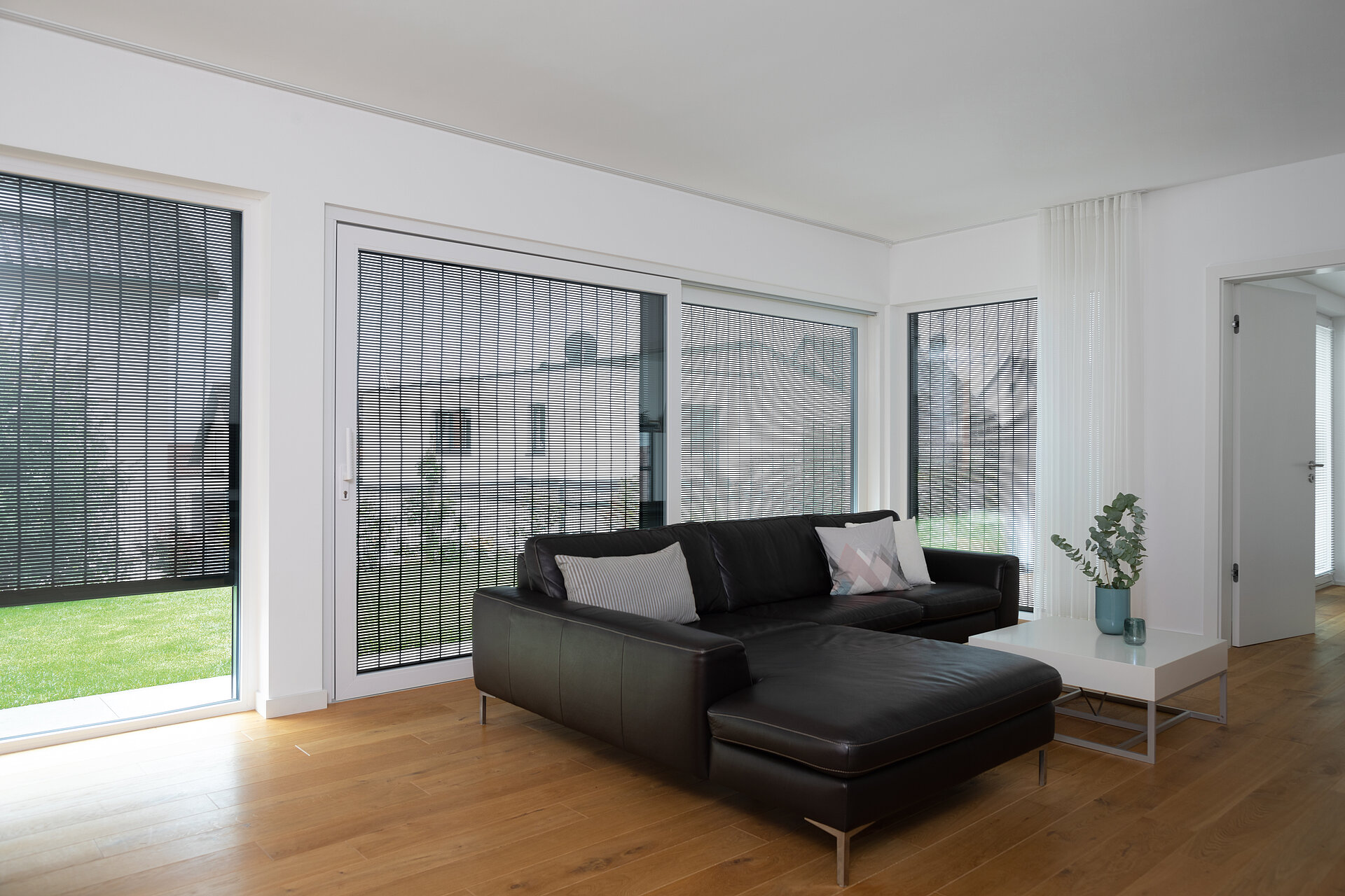Helles Wohnzimmer mit grossem Fenster, schwarzem Ledersofa und MHZ Metallbehang sonro als Sicht- und Sonnenschutz.