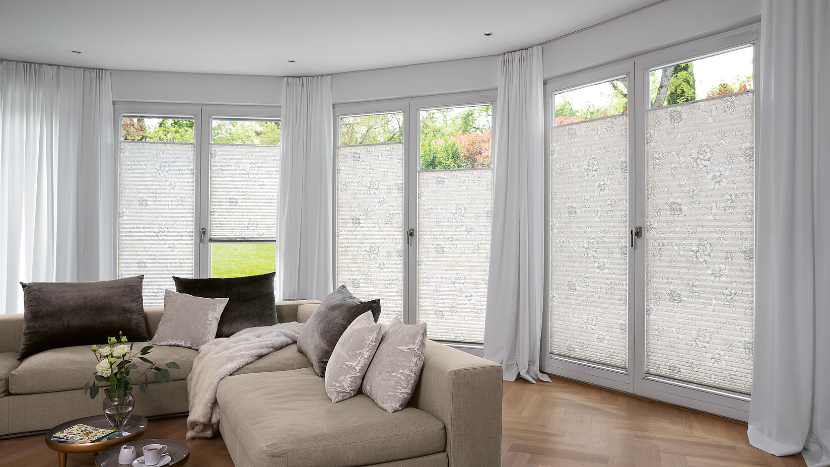 Florale Plissees in Weiss an bodentiefen Fenstern in elegantem Wohnzimmer mit Vorhängen.