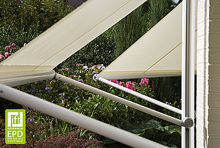 Beige Fallarmmarkisen von MHZ an Fenstern eines Hauses mit Blick auf einen blühenden Garten. Die Markisen sind nach aussen geneigt montiert.