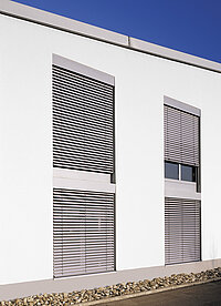 Moderne weisse Fassade mit drei grossen Fenstern, alle mit grauen Jalousien ausgestattet.