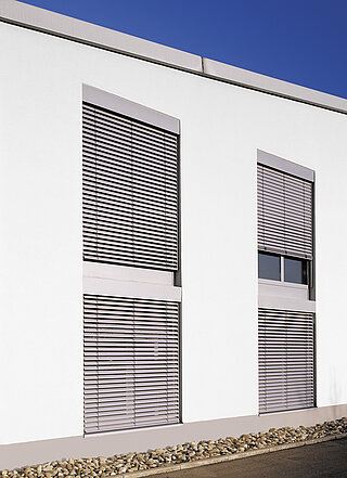 Moderne weisse Fassade mit drei grossen Fenstern, alle mit grauen Jalousien ausgestattet.