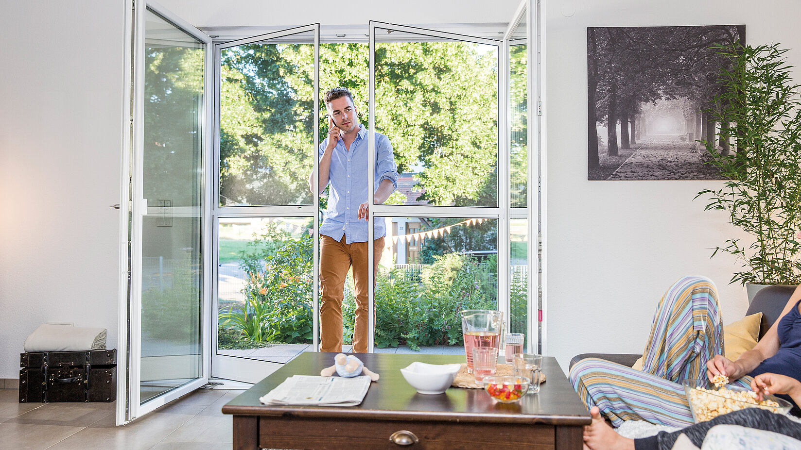 Mann betritt durch eine Pendeltür mit Insektenschutz ein Wohnzimmer, in dem zwei Personen auf dem Sofa sitzen.