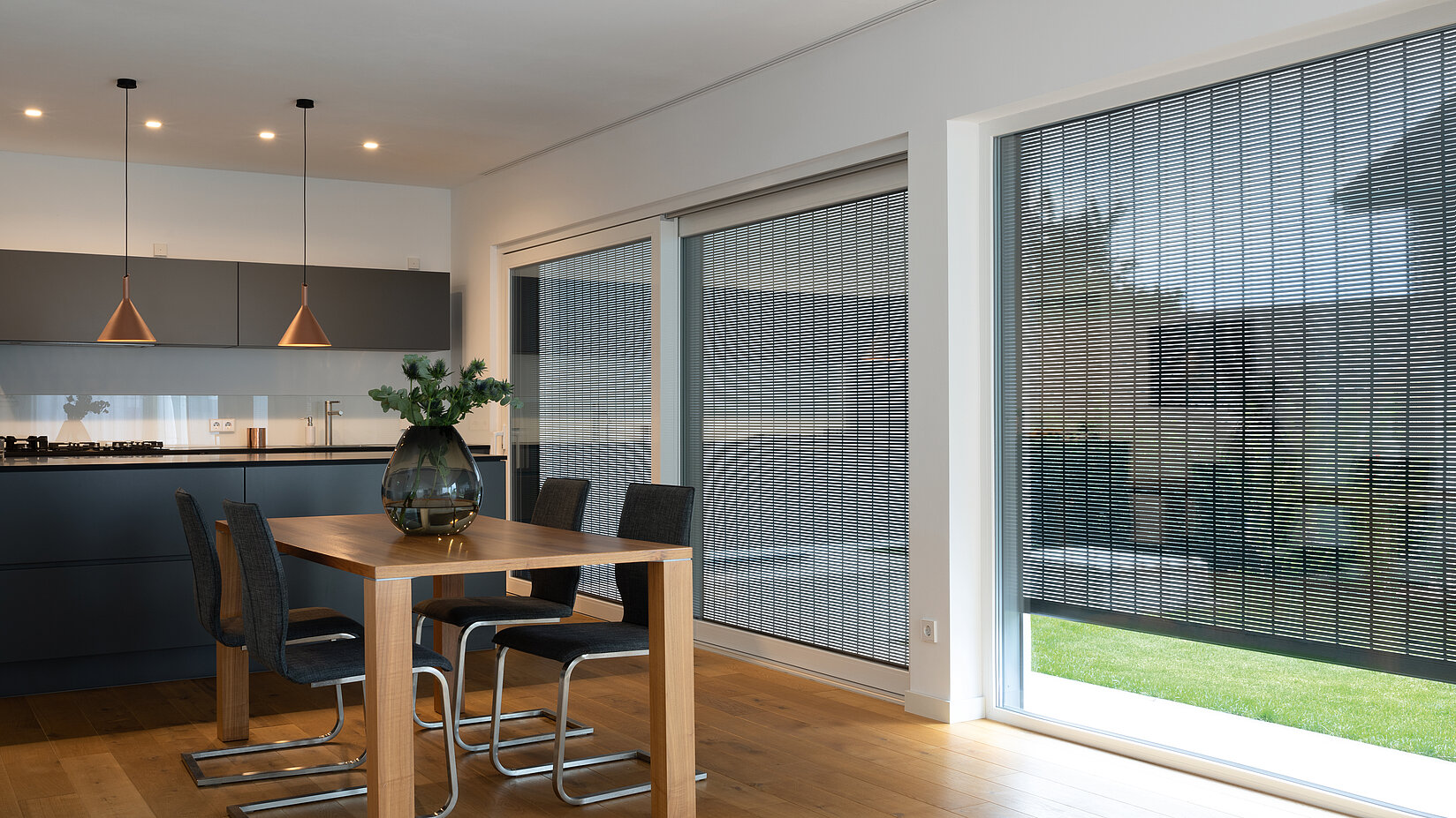 Moderne Wohnküche mit grossen Fenstern und aussenliegendem MHZ Metallbehang Sonro, Holztisch im Vordergrund.