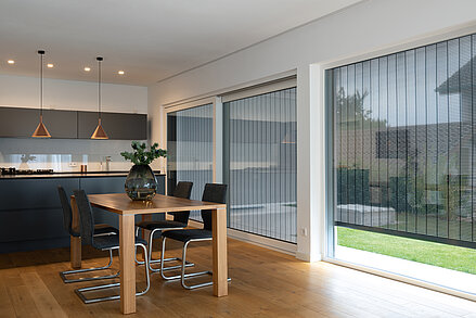 Moderne Wohnküche mit grossen Fenstern und aussenliegendem MHZ Metallbehang Sonro, Holztisch im Vordergrund.