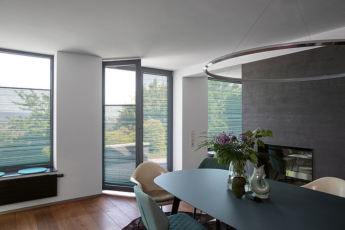 Petrolfarbene Plissees an bodentiefen Fenstern im modernen Esszimmer mit grünem Tisch.