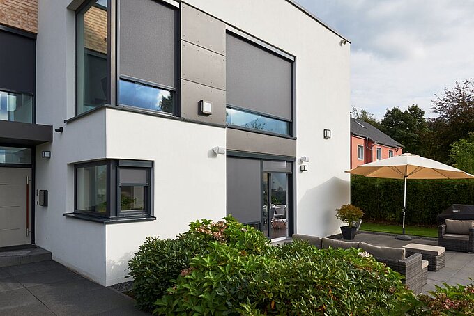 Modernes Wohnhaus mit geschlossenen Senkrechtmarkisen an grossen Fenstern und Terrasse mit Sonnenschirm.
