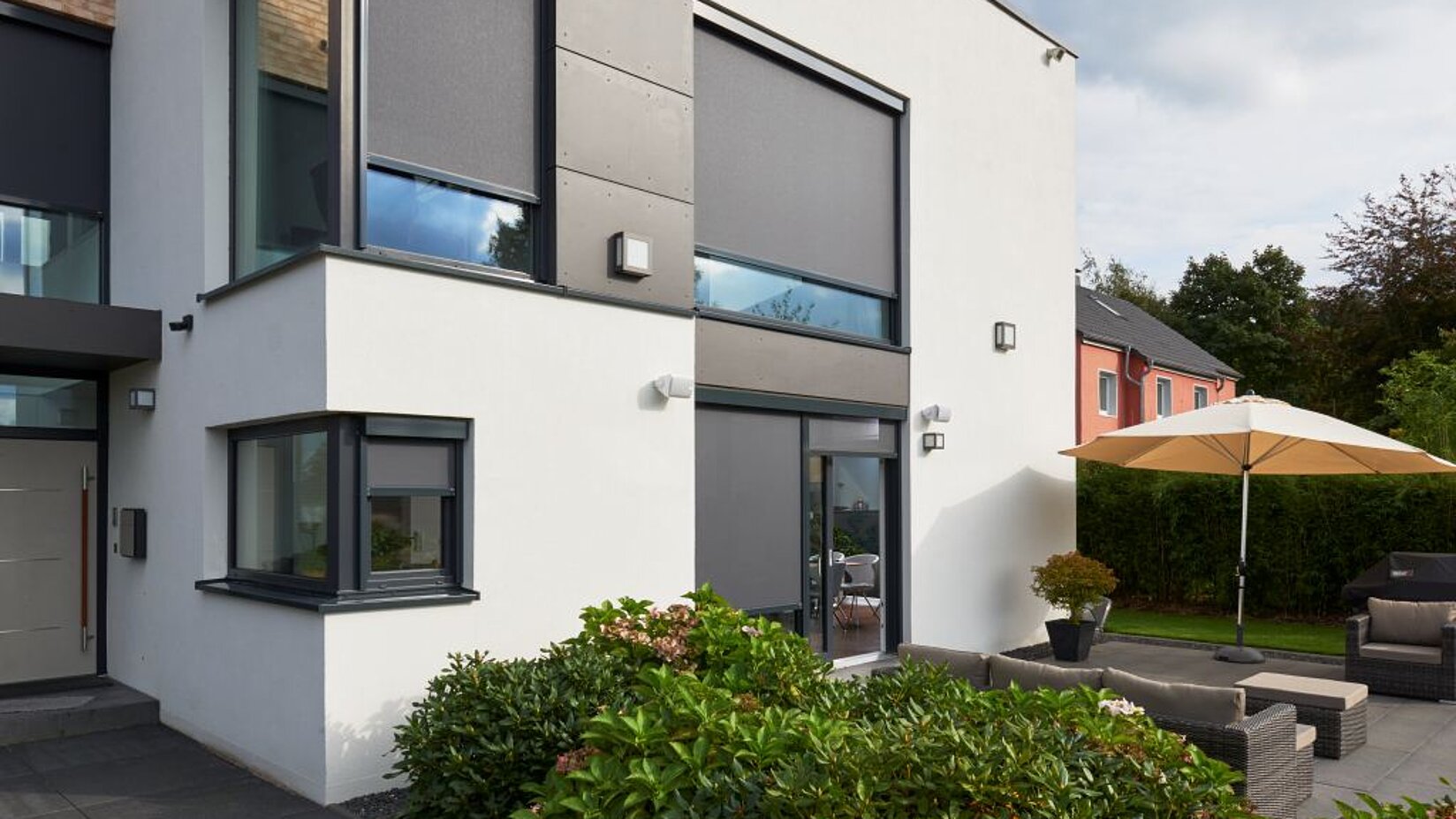 Modernes Wohnhaus mit geschlossenen Senkrechtmarkisen an grossen Fenstern und Terrasse mit Sonnenschirm.