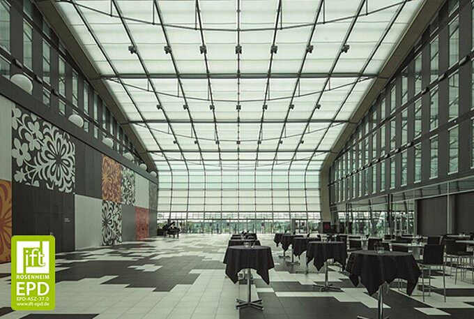 Ein lichtdurchfluteter Innenraum mit hoher Glasdecke, die durch das MHZ-KOAX-Beschattungssystem mit textilen Flächen verschattet ist. Der Raum ist mit Tischen und Stühlen bestückt.