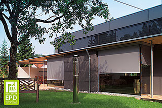 Ein modernes Wohnhaus mit grossen Fenstern, die durch aussenliegende Kassettenrollos von MHZ verschattet werden. Im Vordergrund befindet sich ein Garten mit Bäumen und Rasenfläche.