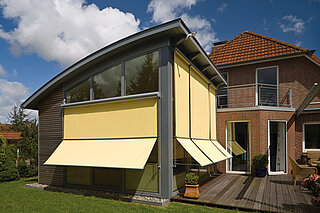 Modernes Wohnhaus mit gelben ZIP-Senkrechtmarkisen an grossen Fenstern und Holzterrasse im Garten.